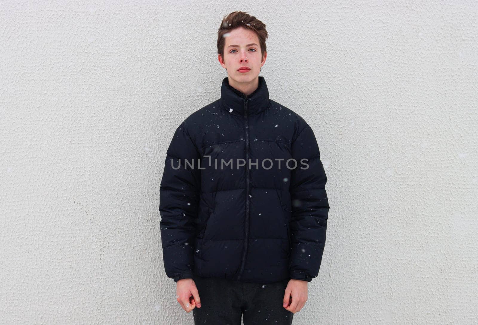 A belted portrait of a teenage boy in a black jacket against a wall in wintertime. A boy against a background of falling snowflakes.