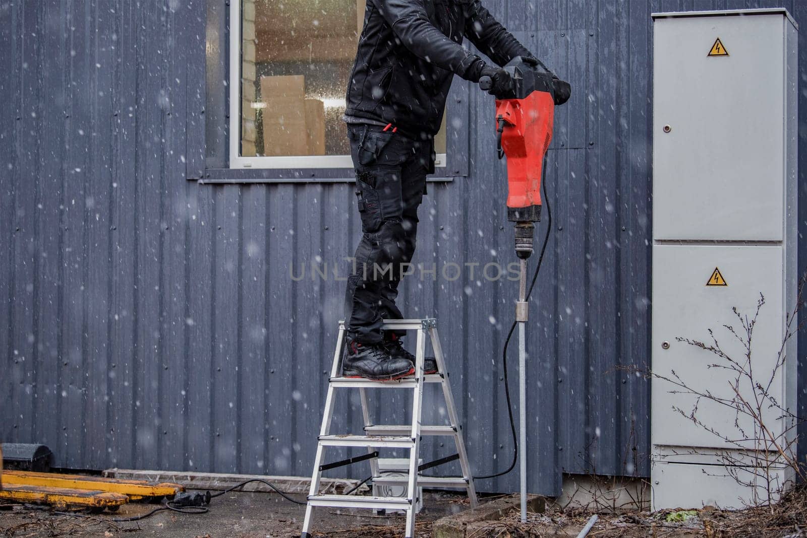 A worker installs a ground rod to ground a building. A worker in work clothes drives earth rods into the ground with a jackhammer to prevent short circuits. by SERSOL