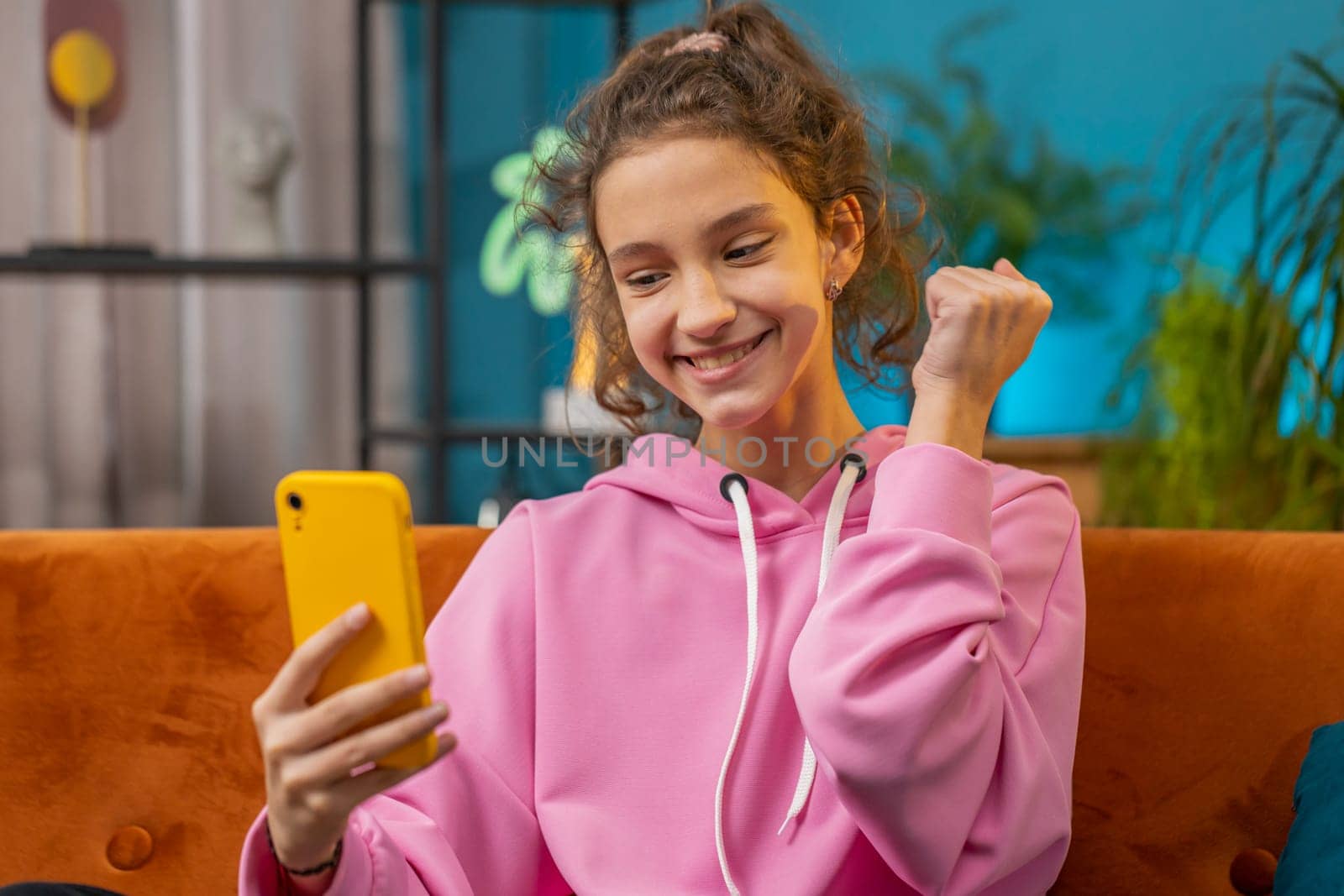 Preteen school girl use mobile smartphone celebrate success win triumph victory, positive test, exam results, play game. Young child kid gets online good news at home room apartment sitting on couch
