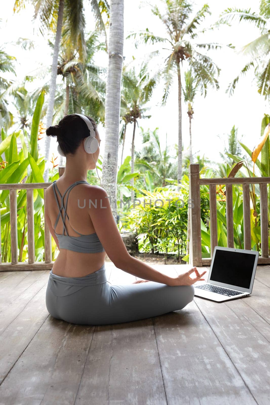Young woman in sports clothing following online meditation using laptop and headphones outdoors. Vertical image. Spirituality and mental health concept.