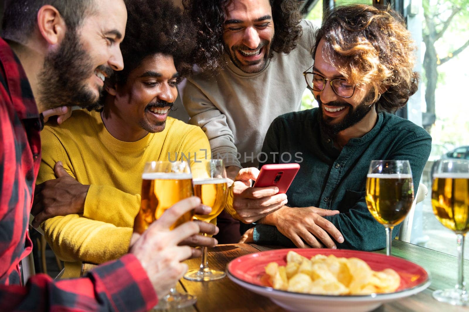 Multiracial guy friends looking at mobile phone together while having drinks together in pub. Vertical image. Technology and leisure activity.