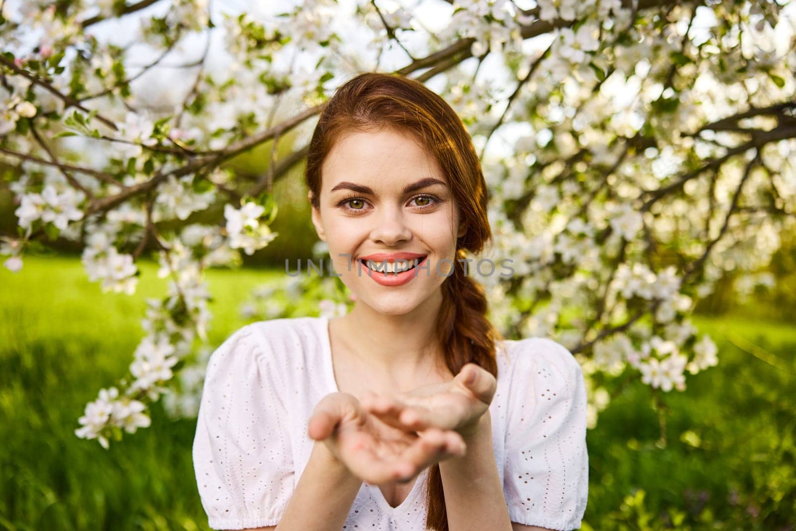 openly smiling woman against the backdrop of a flowering tree stretches her palms to the camera by Vichizh