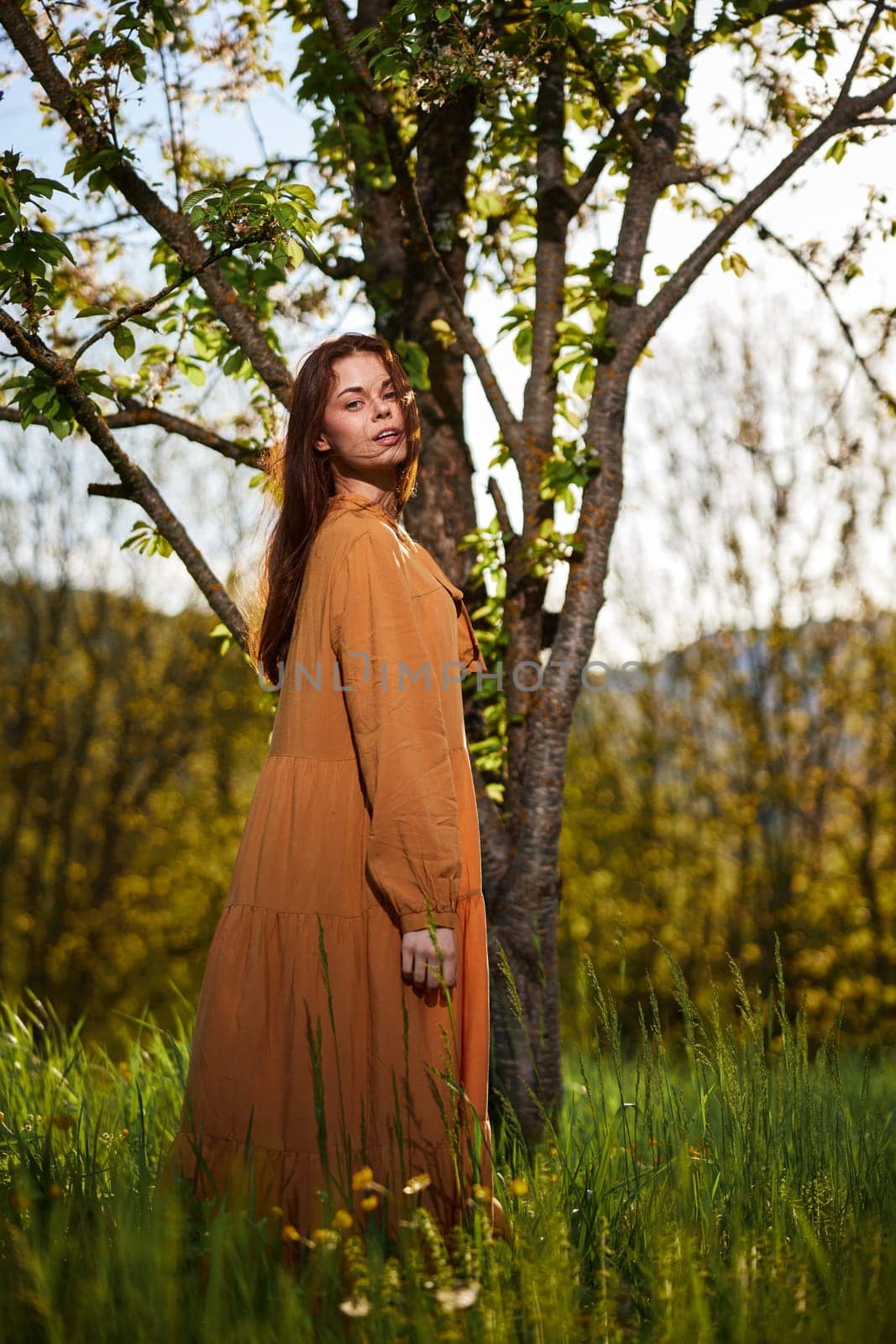 a sweet, attractive woman with long red hair stands in the countryside near a flowering tree in a long orange dress and looks at the camera with a slight smile on her face. High quality photo
