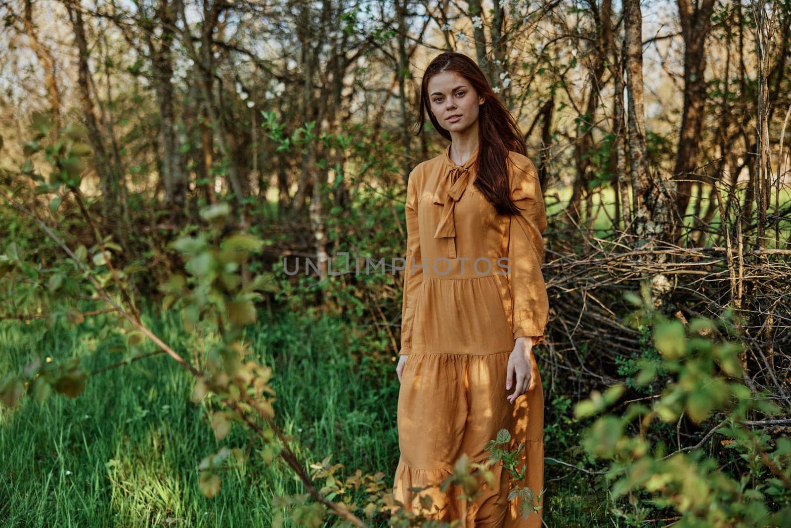 a woman with long hair walks in the shade near the trees, dressed in a long orange dress, enjoying the weather and the weekend, while looking at the camera. The theme of privacy with nature. High quality photo