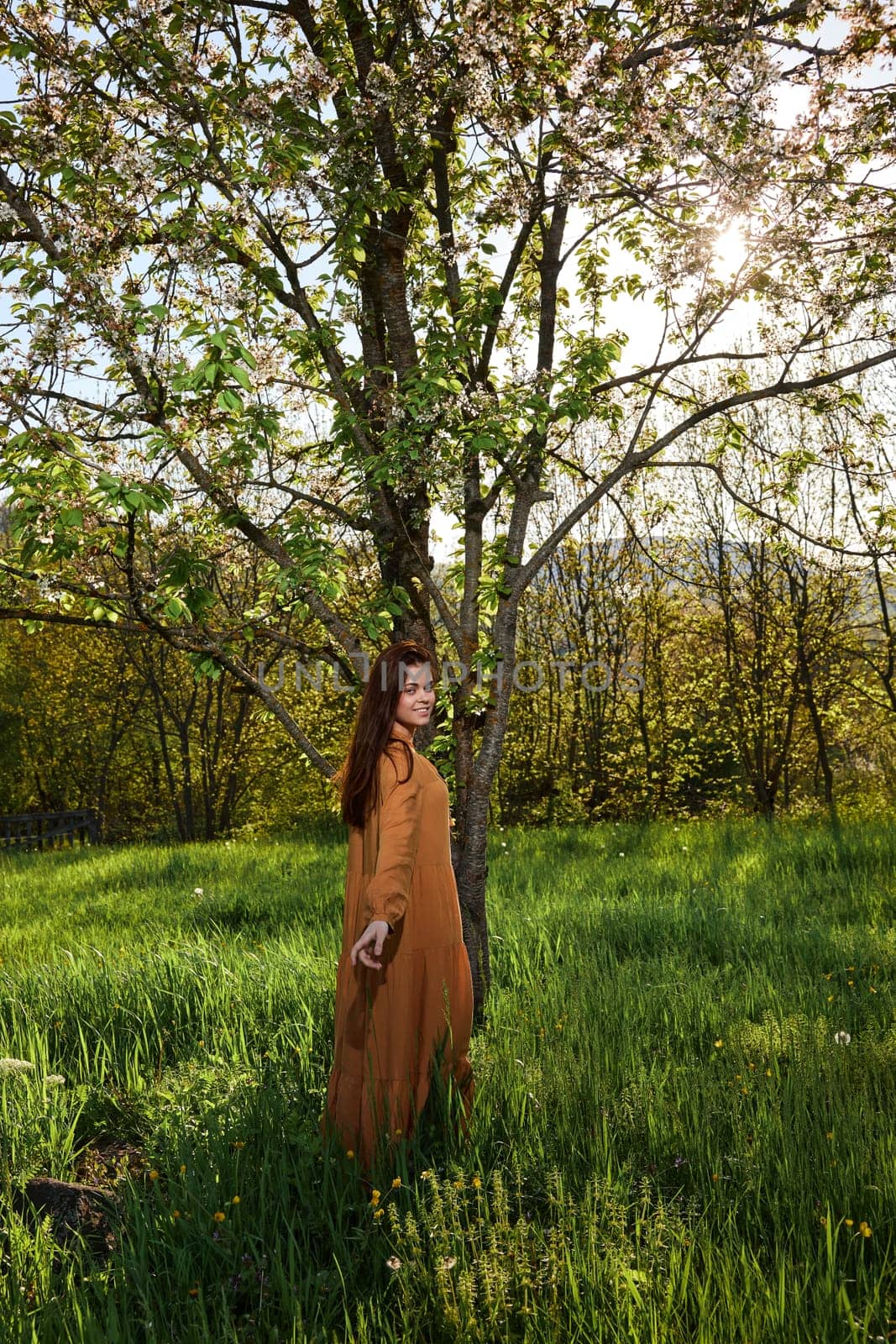 a slender, calm woman with long red hair stands in the countryside near a flowering tree in a long orange dress and looks into the camera enjoying the silence. High quality photo