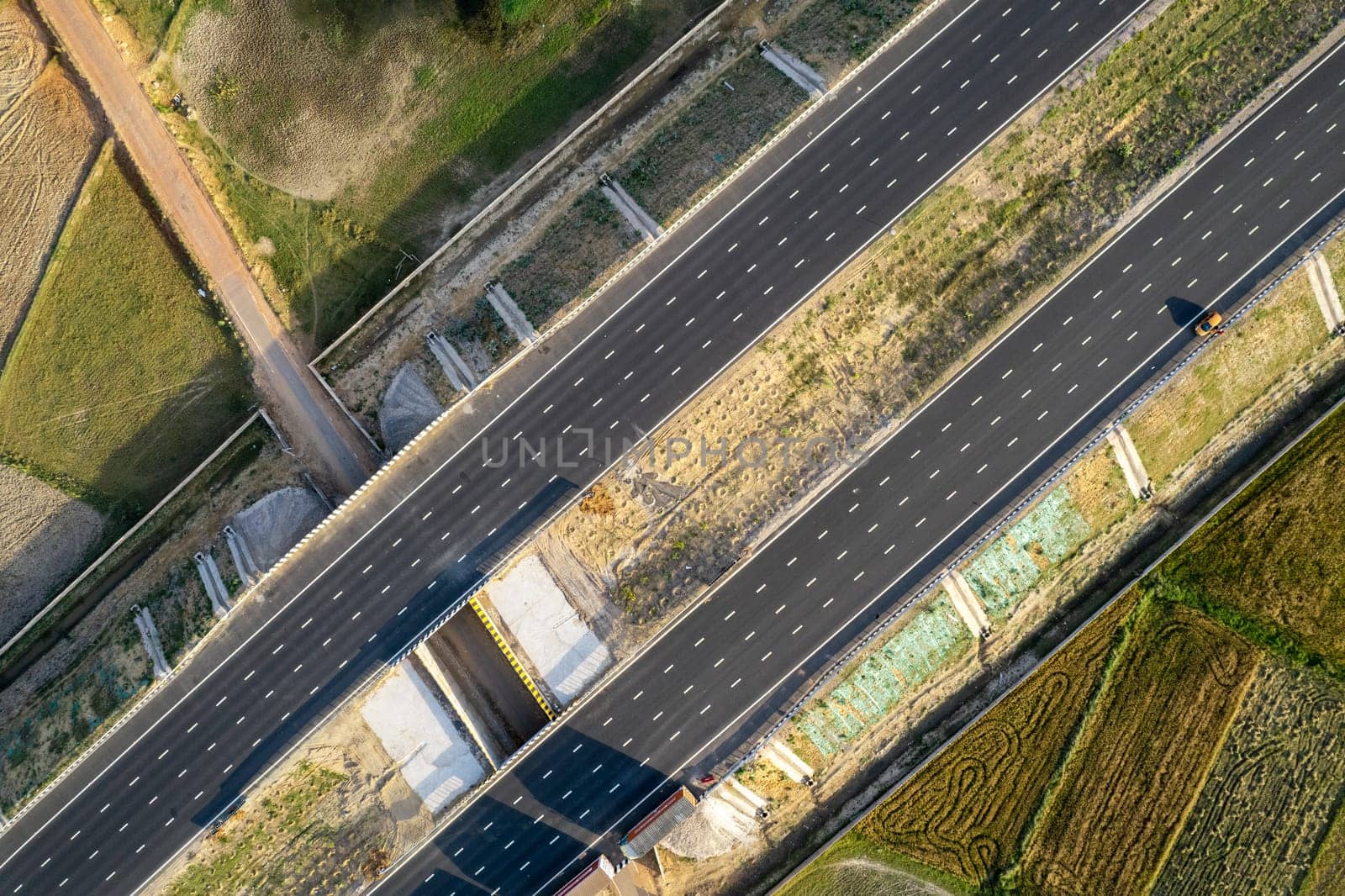 aerial drone shot moving along and shooting straight down of new delhi mumbai jaipur express elevated highway showing six lane road with green feilds with rectangular farm by Shalinimathur