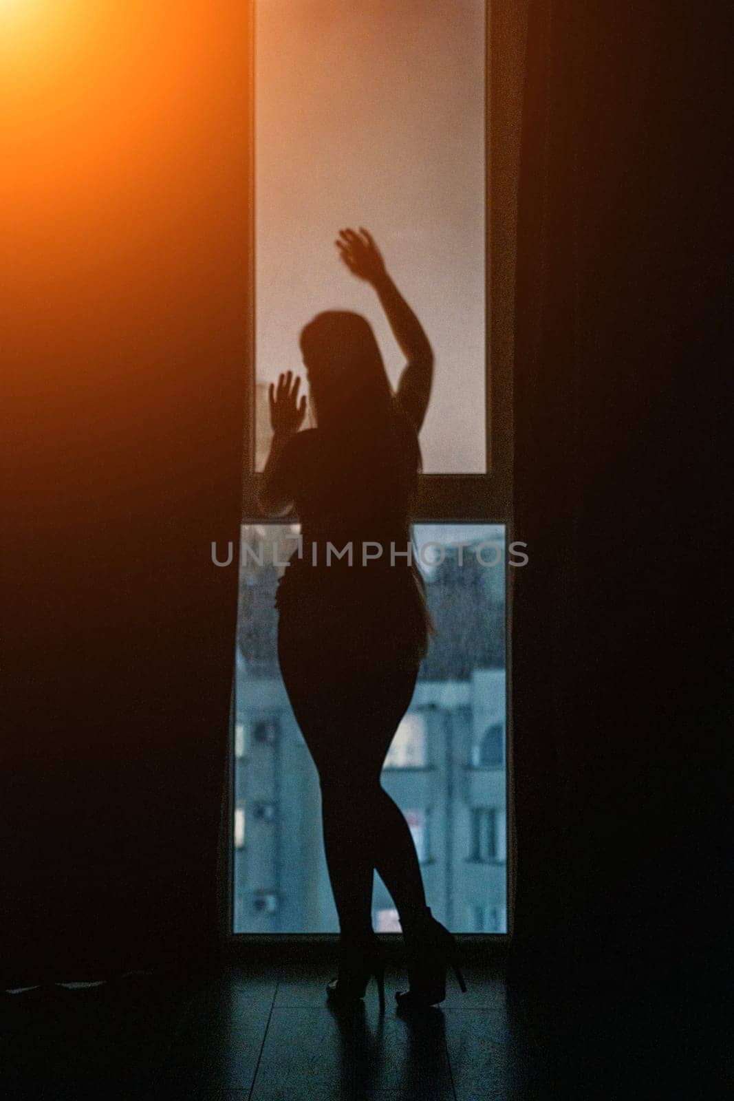 A full-length silhouette of young woman at open balcony window and dark blue night sky, with a cityscape in the background. Seduction and passion concept. Romantic moment in the bedroom. Night room