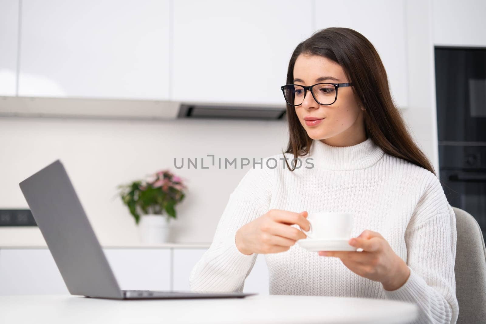 Confident young woman with long dark hair and glasses sitting at table with opened laptop and drinking coffee. SMM manager working at home.