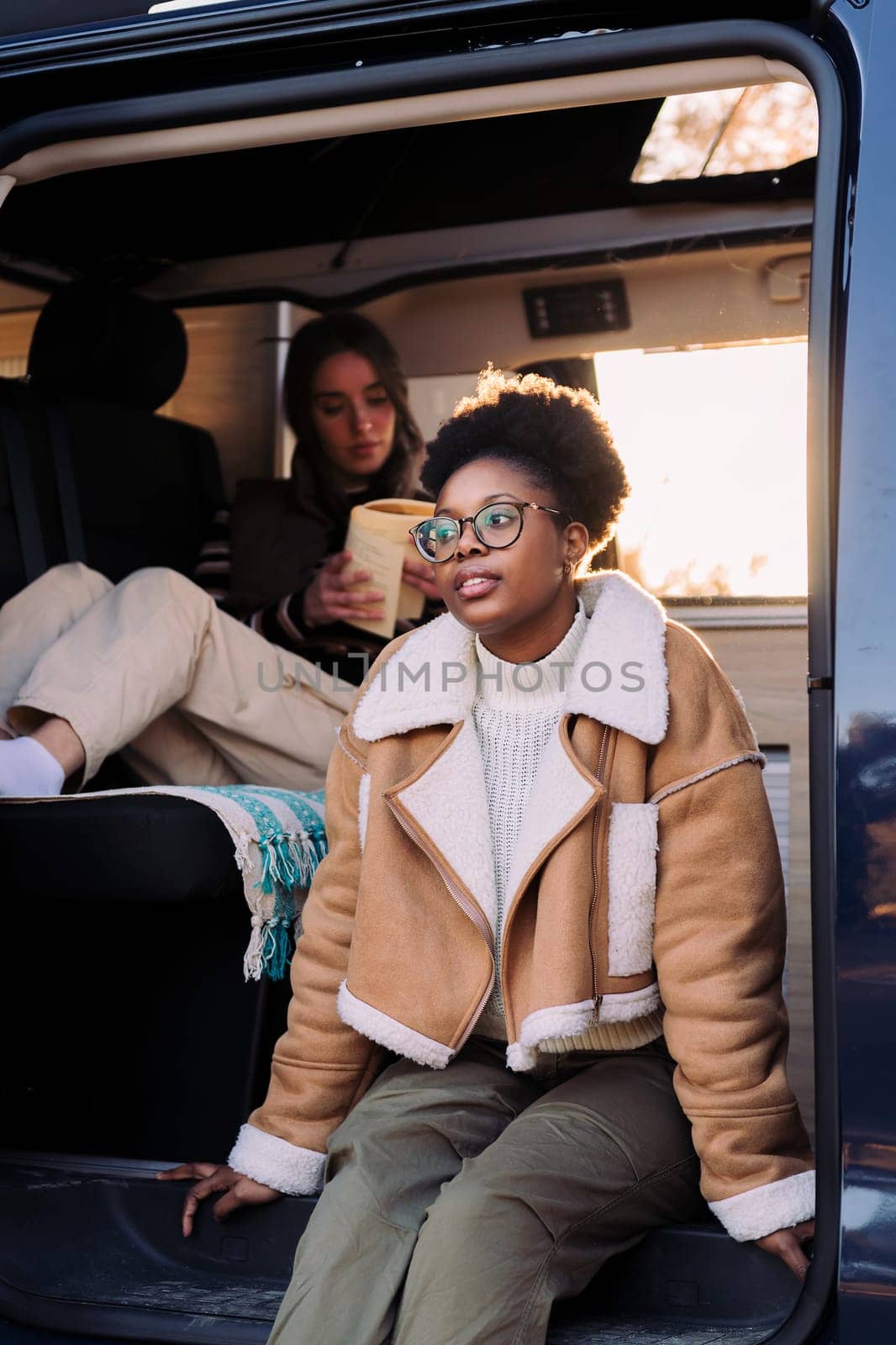 young african woman sitting at sunset in a camper van with a friend reading in the background, concept of travel adventure and female friendship