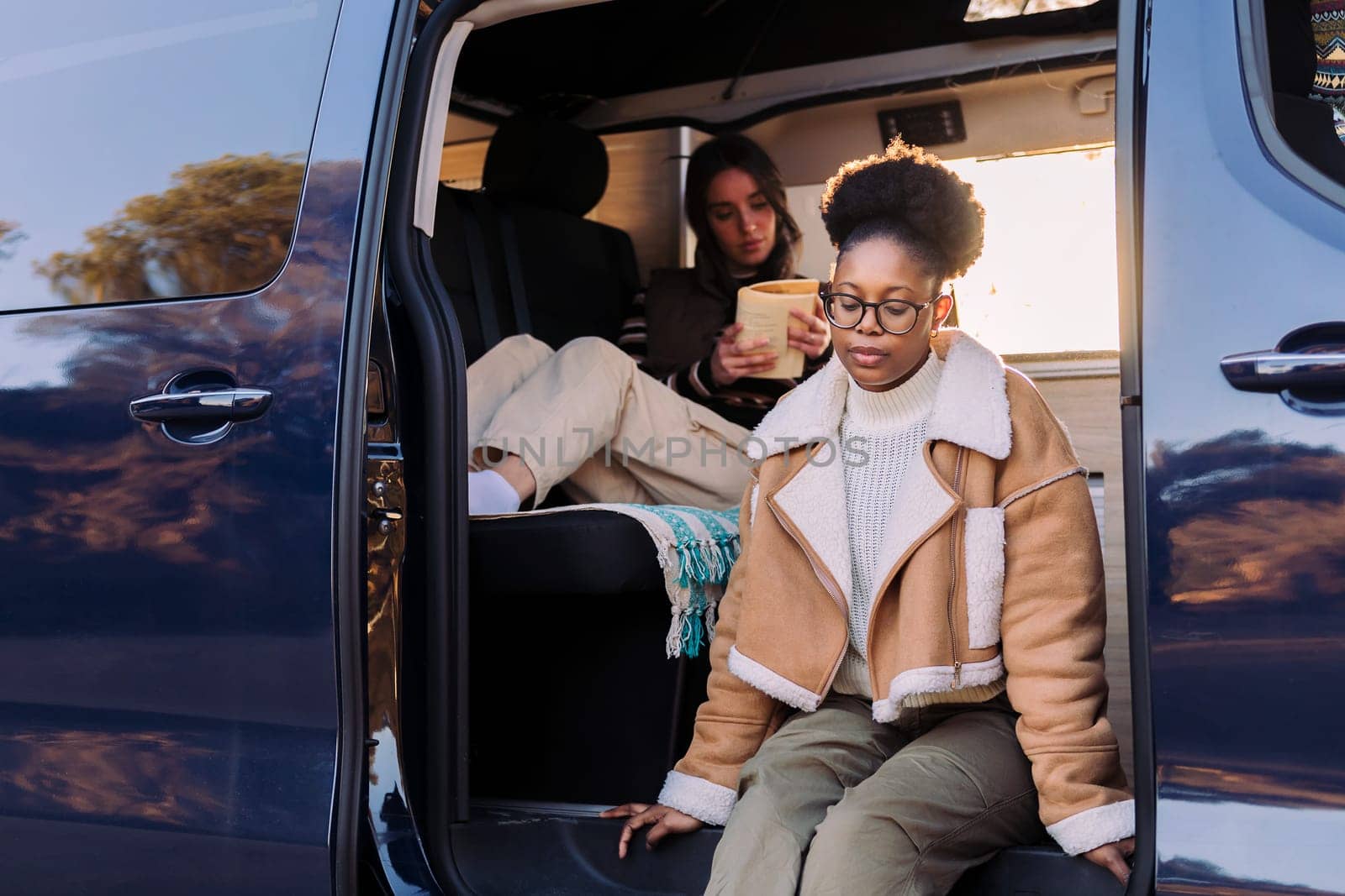 young african woman sitting at sunset in a camper van with a friend reading in the background, concept of travel adventure and female friendship
