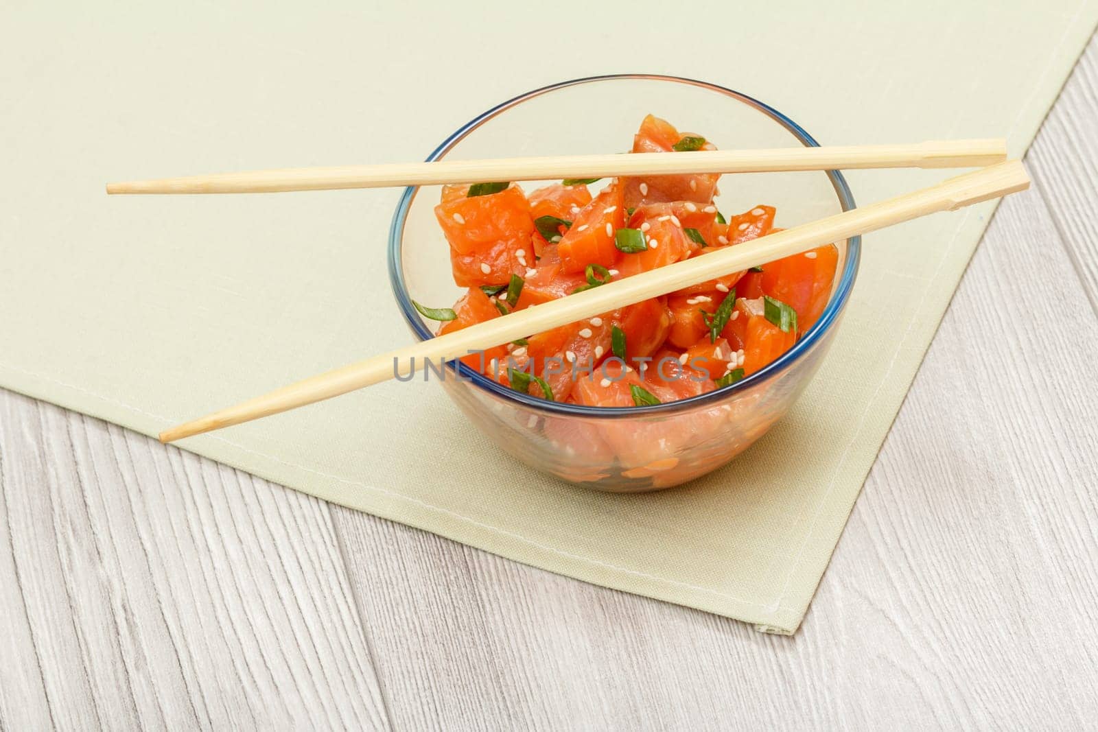 Hawaiian salmon poke with green onions and sesame seeds in glass bowl with chopsticks on kitchen napkin. Top view. Organic seafood.