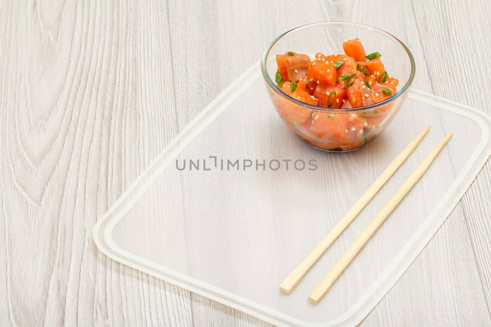 Hawaiian salmon poke with green onions and sesame seeds in glass bowl with chopsticks on kitchen napkin. Top view with copy space. Organic seafood.