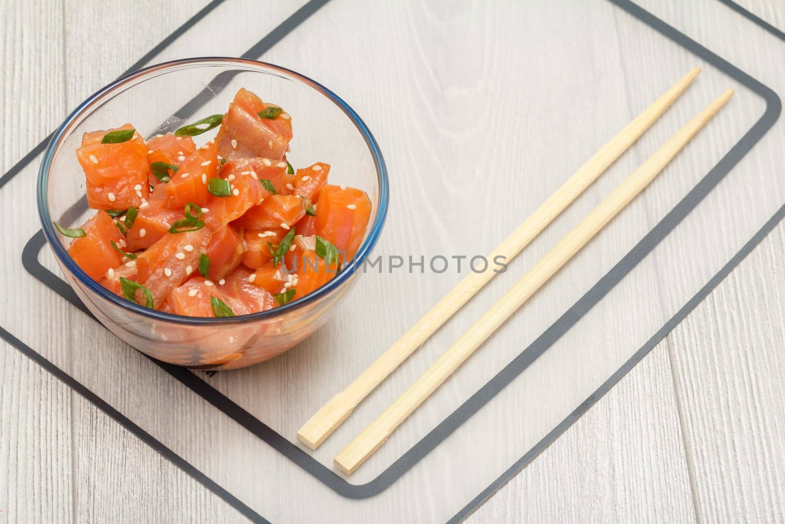 Hawaiian salmon poke with green onions and sesame seeds in glass bowl with chopsticks on wooden background. Top view. Organic seafood.
