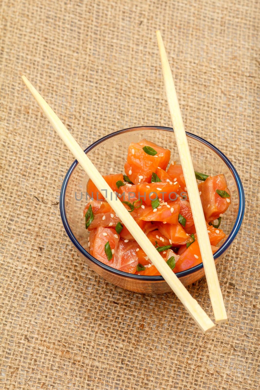 Hawaiian salmon poke with green onions and sesame seeds in glass bowl with chopsticks on sackcloth. Top view. Organic seafood.