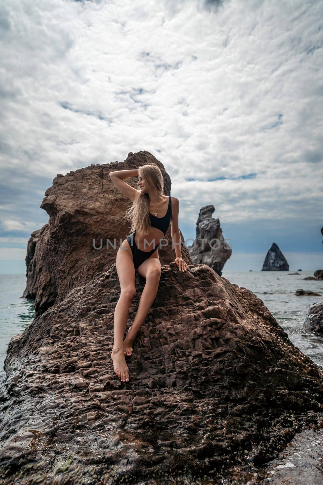 Woman swimsuit sea. Attractive blonde woman in a black swimsuit enjoying the sea air on the seashore around the rocks. Travel and vacation concept. by Matiunina