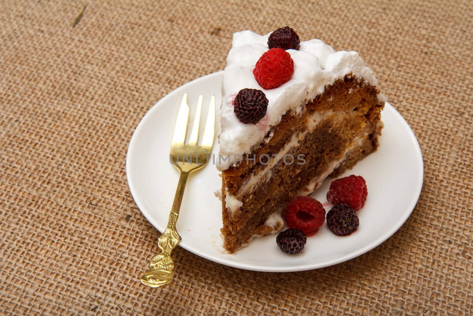 Slice of homemade biscuit cake decorated with whipped cream and raspberries on plate with sackcloth background.
