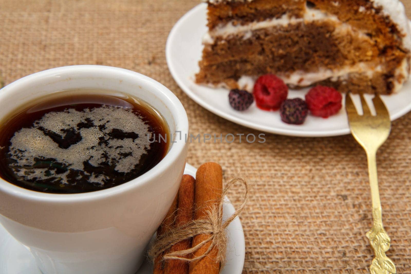 Cup of coffee, cinnamon, fork and slice of biscuit cake decorated with whipped cream and raspberries on table with sackcloth.
