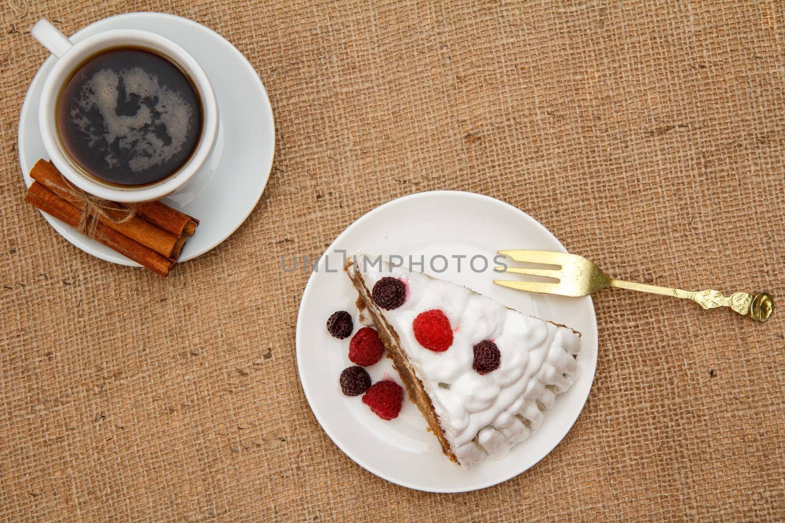 Cup of coffee, cinnamon, fork and slice of biscuit cake decorated with whipped cream and raspberries on table with sackcloth. Top view.