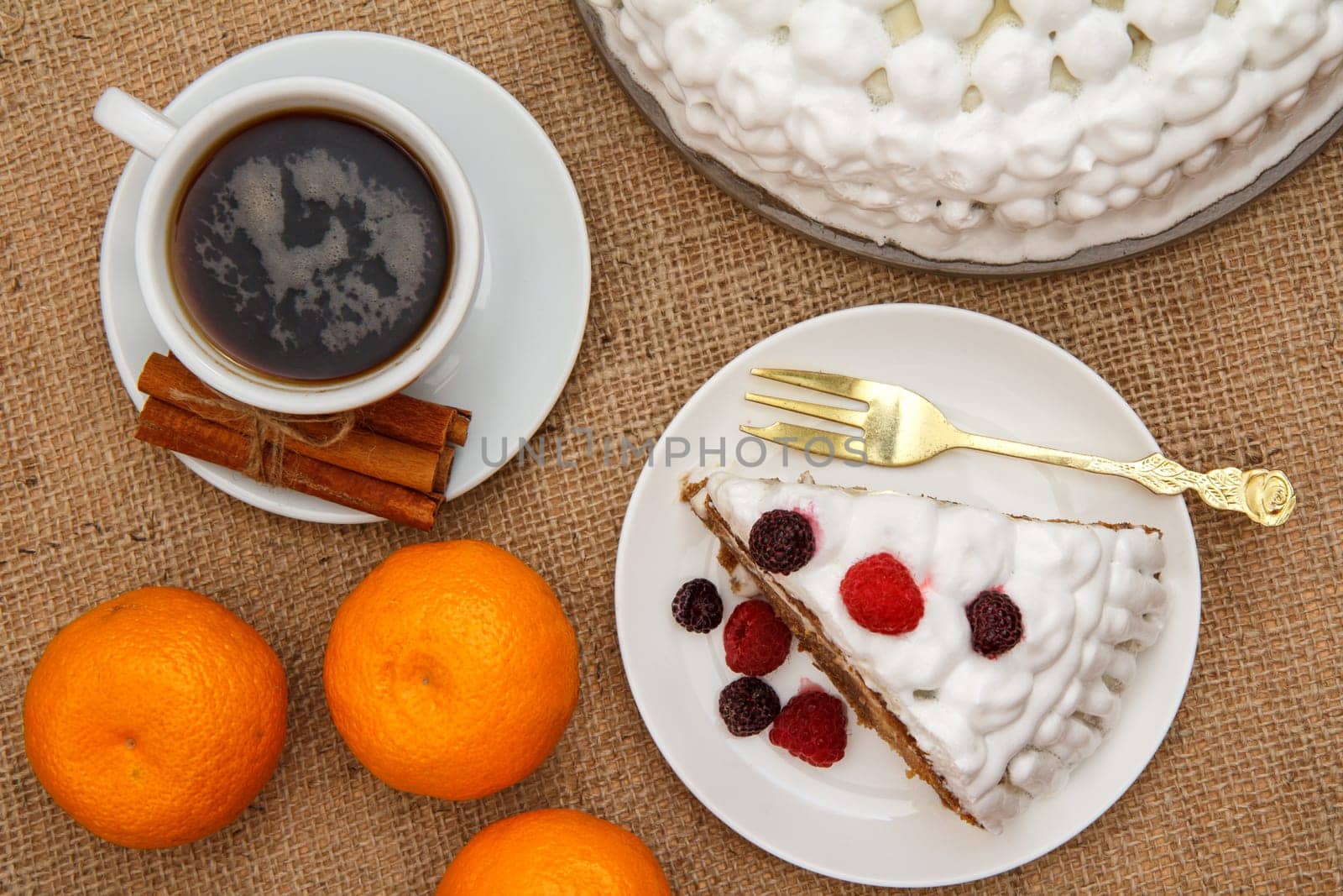 Cup of coffee, cinnamon, fork and slice of biscuit cake decorated with whipped cream and raspberries, oranges on table with sackcloth. Top view.