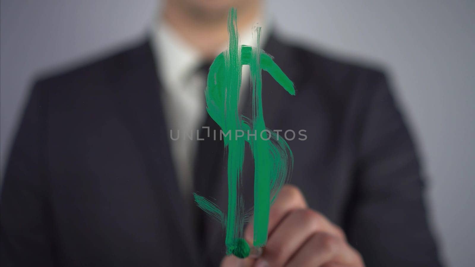 A young businessman draws a dollar symbol on a transparent glass. A man draws with green paint and a brush close-up. 4k