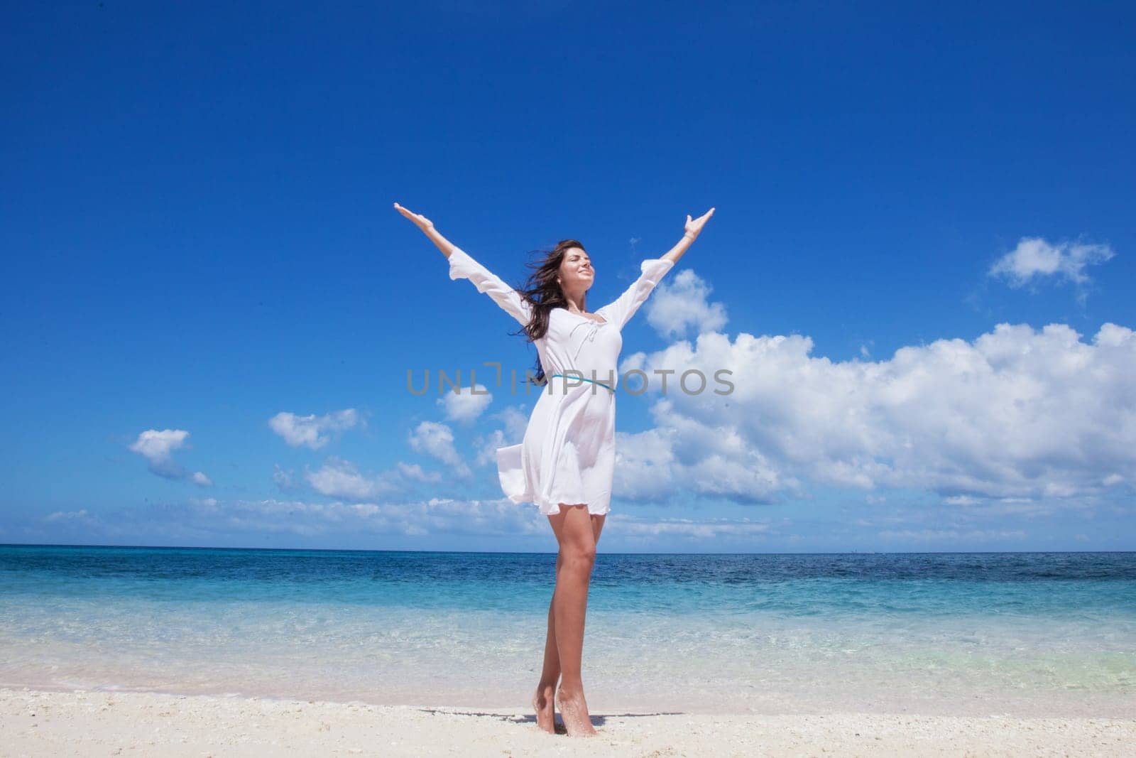 Woman in white dress posing in tropical sea beach with arms raised