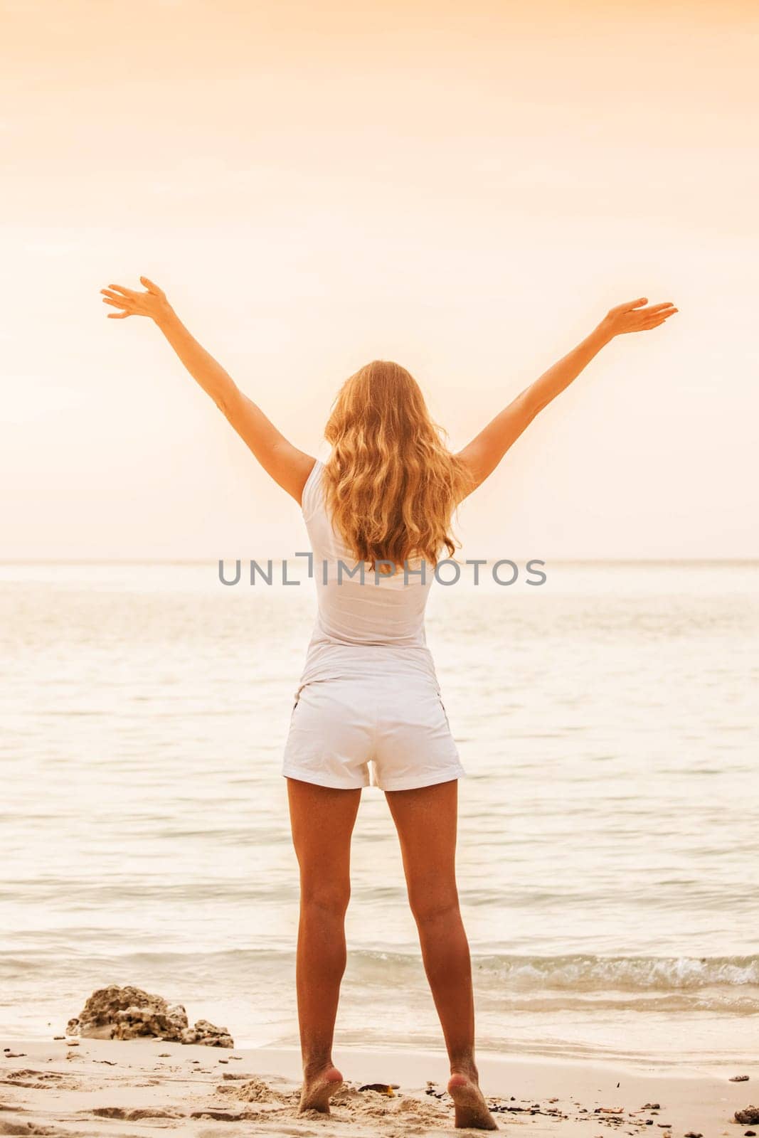 Beautiful young woman standing on sandy beach against orange sky at sunset. Summer travel. Happy slim girl in white with raised up arms on the seashore