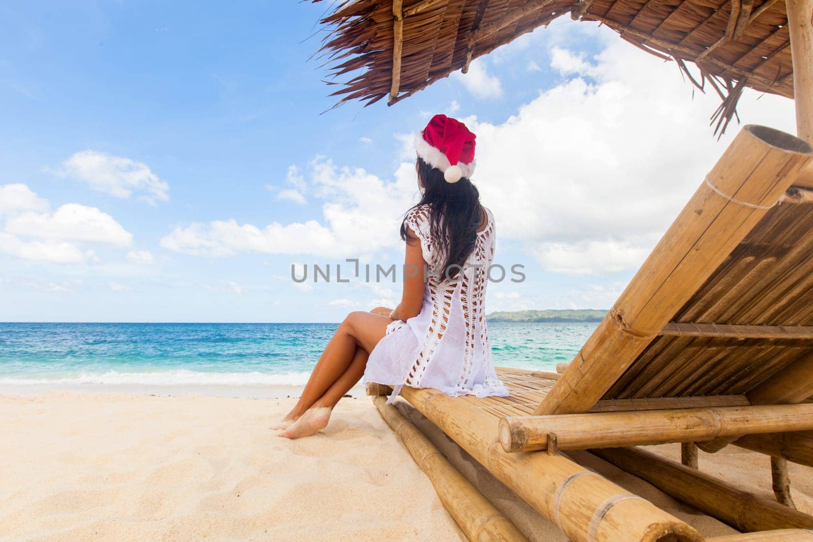 Woman in Santa hat sitting at chaise lounge with straw parasol on white sandy beach at Philippines