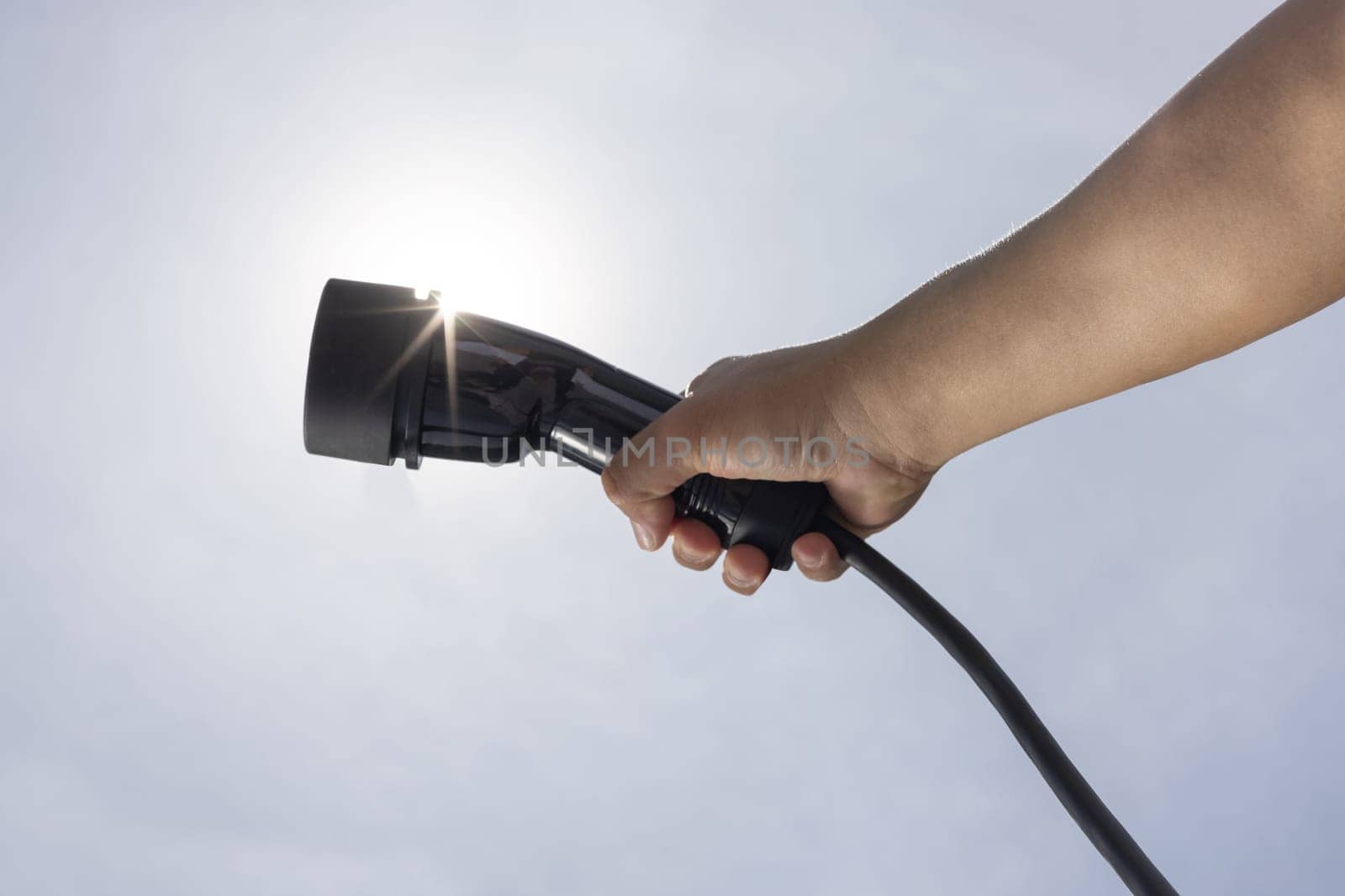 Hand holding EV charger device with the view from below showing sky, cloudscape and sun in the background as concept for progressive sustainable energy for eco-friendly electric car.