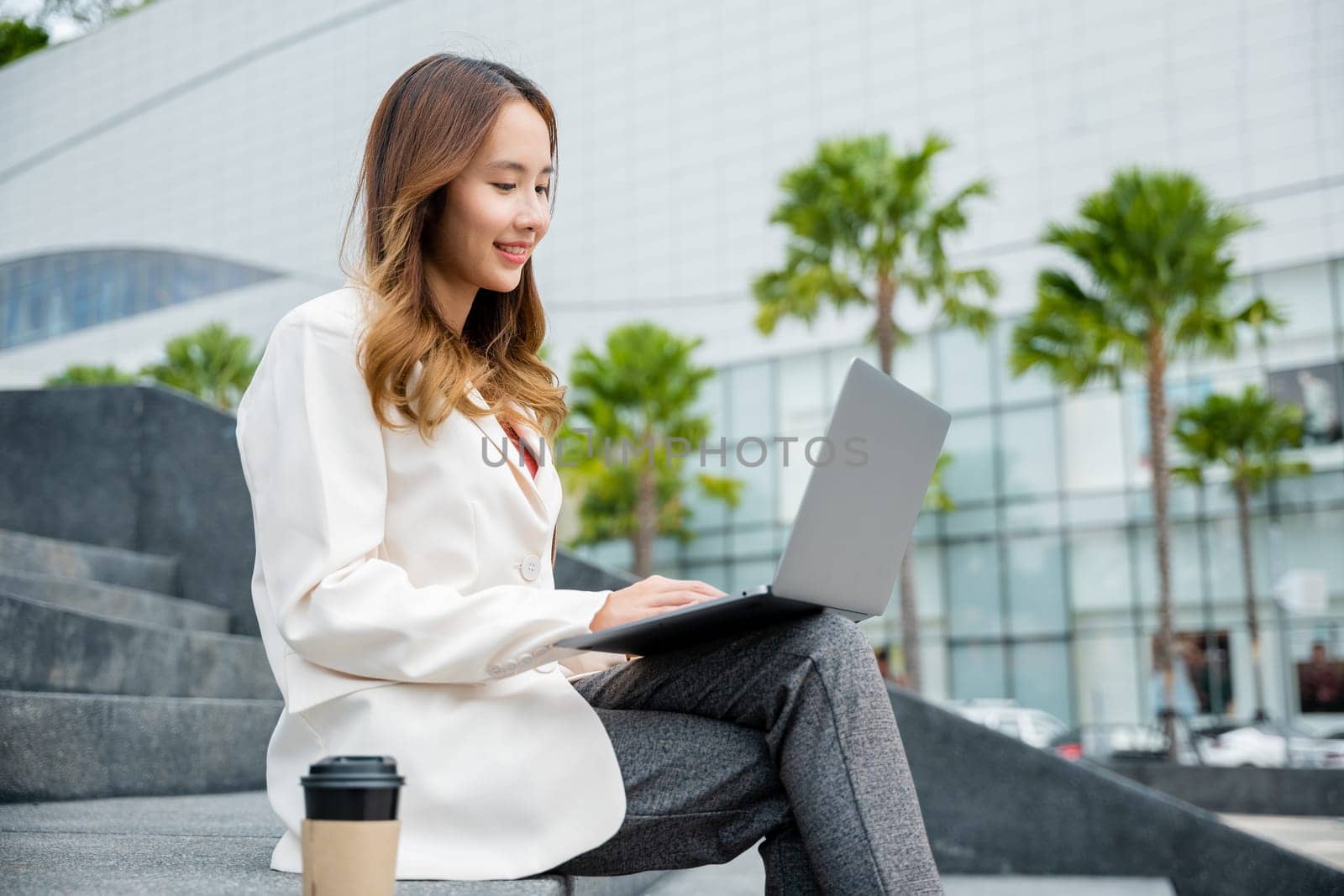 Happy young Asian woman freelance looking laptop with smile while working with coffee cup while sitting on stairs outdoors in the city. Remote working concept in the office.