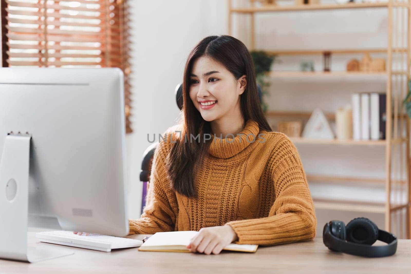 Online education, e-learning. Asian woman in stylish casual clothes, studying using a computer, listening to online lecture, taking notes, online study at home.