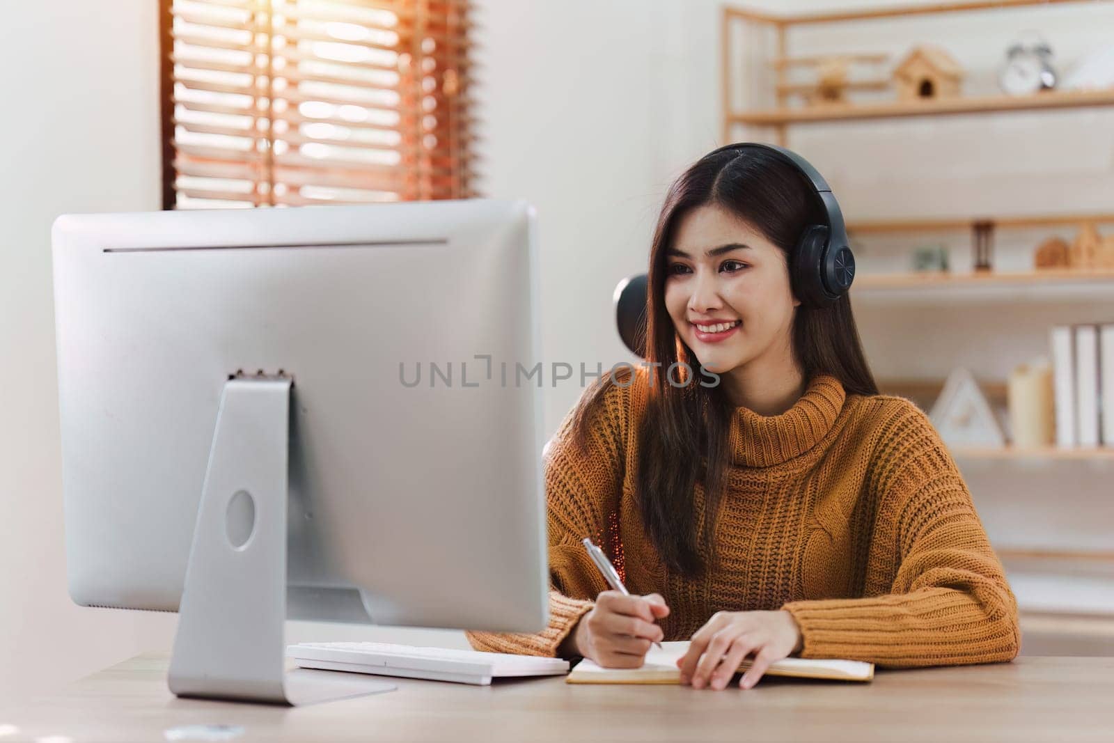 Online education, e-learning. Asian woman in stylish casual clothes, studying using a computer, listening to online lecture, taking notes, online study at home.