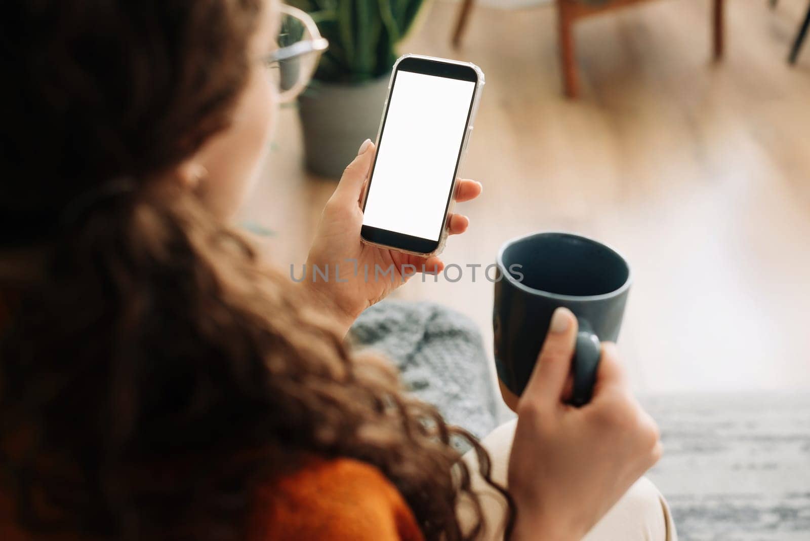 Over-shoulder view of woman holding coffee and smartphone with white mock-up cellular screen in hands using mobile apps on a cell phone. Cellphone display mock-up for advertising concept