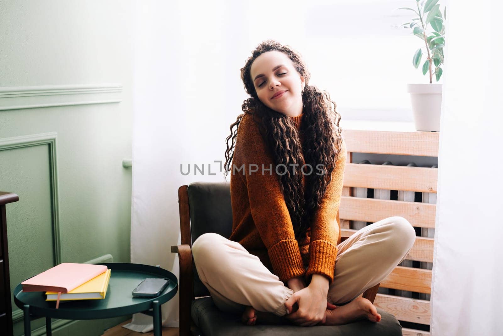 Young relaxed smiling pretty woman relaxing sitting on chair at home. Happy positive beautiful lady feeling joy enjoying wellbeing and lounge chilling near window in modern cozy apartment interior