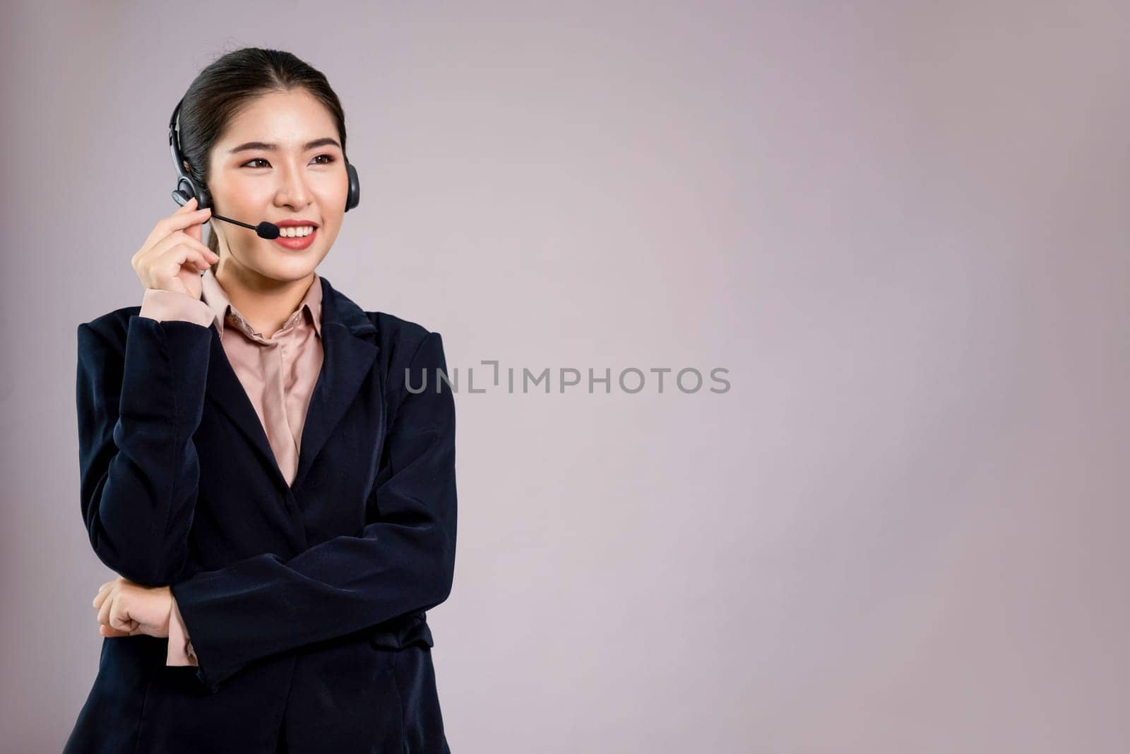 Attractive asian female call center operator with happy smile face advertises job opportunity on empty space, wearing a formal suit and headset on customizable isolated background. Enthusiastic