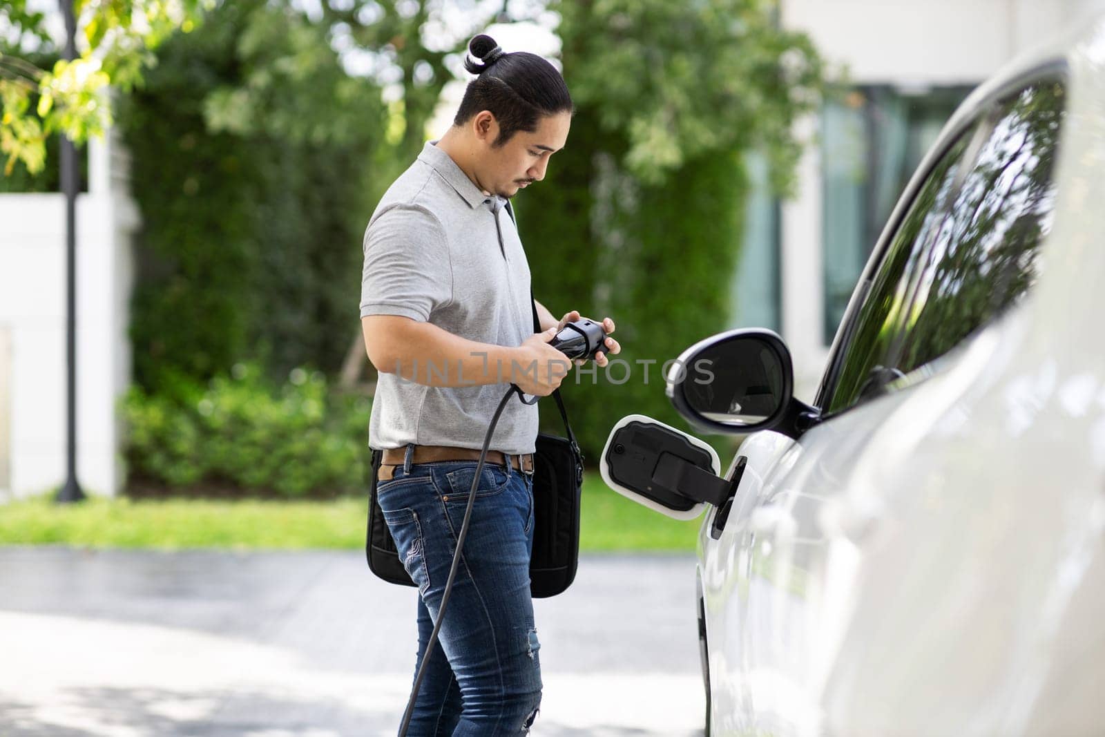 Progressive asian man install cable plug to his electric car with home charging station in the backyard. Concept use of electric vehicles in a progressive lifestyle contributes to clean environment.