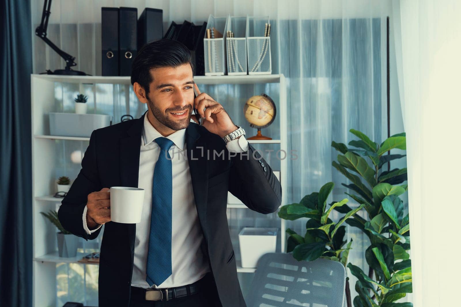 Diligent and busy businessman talking on phone call with coworker in modern office. Making sales call to client with coffee in hand, standing posing confidently as professional office worker. Fervent