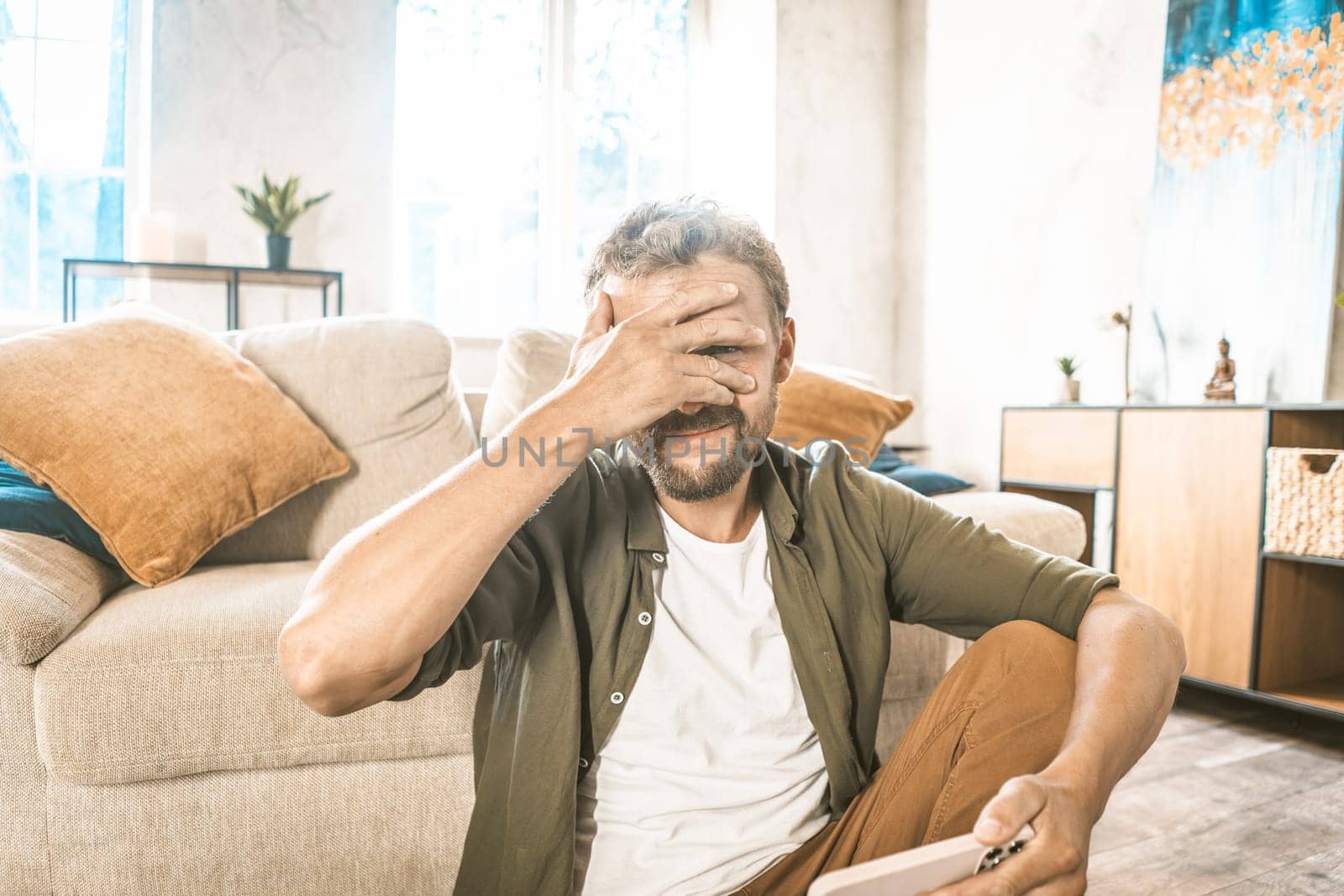 Cheater online. Man covering face with facepalm, holding mobile phone. Hacker concept. He may have been caught doing something wrong or possibly caught in act of hacking or online fraud. by LipikStockMedia