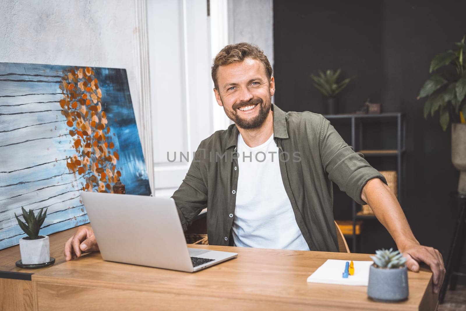 Concept of remote work, freelance, and productivity in digital age. Cheerful and productive man, works from home, He smiling, focusing on his work, surrounded by digital technology and equipment. by LipikStockMedia