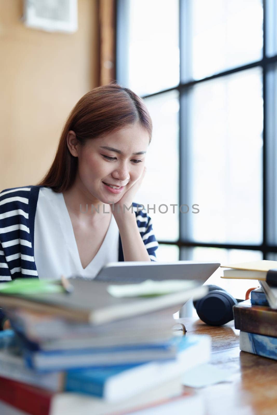 A portrait of a young Asian woman with a smiling face using a tablet computer during an online video conferencing class in a library.