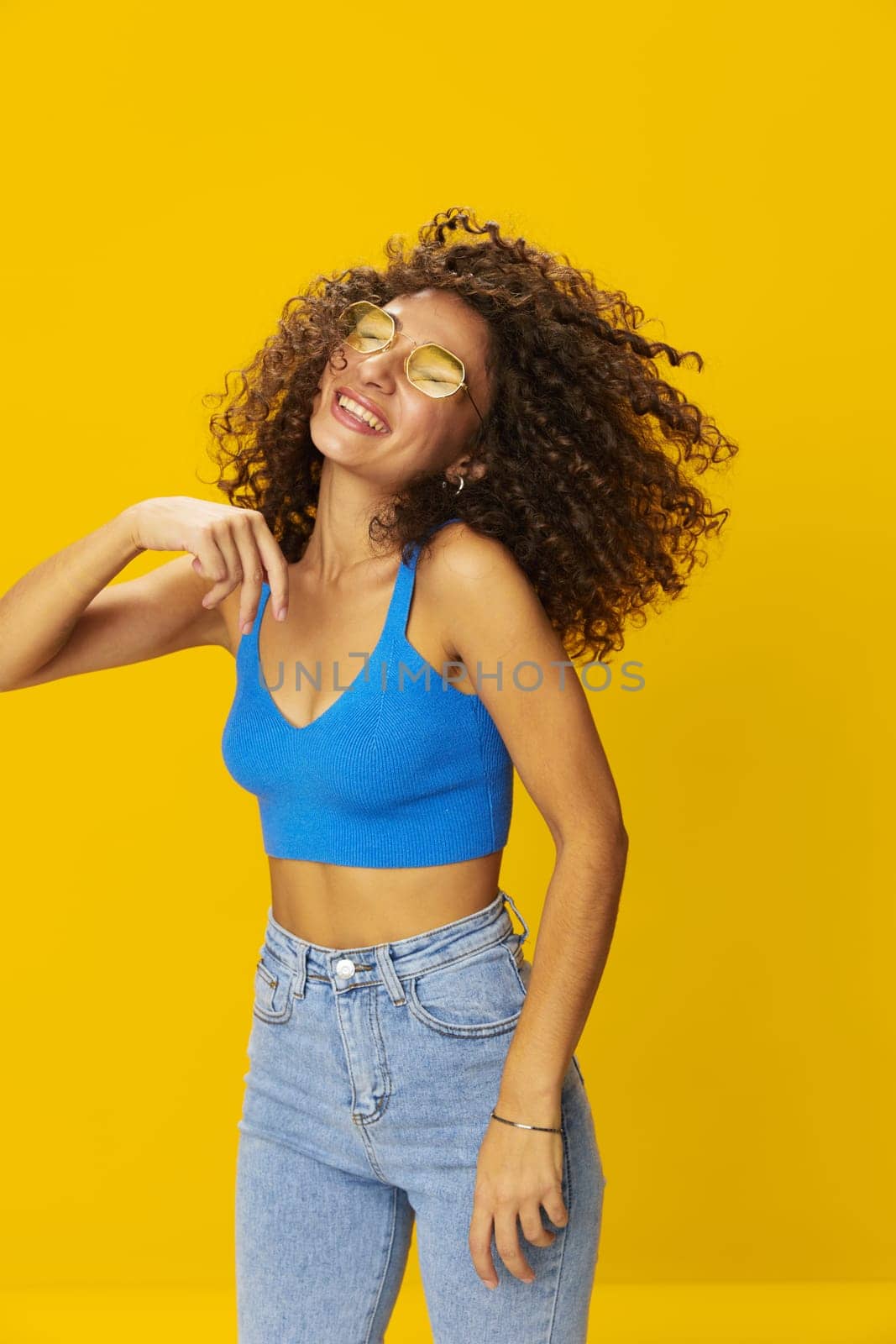 Woman with curly afro hair in a blue T-shirt on a yellow background dancing flying hair with sunglasses yellow, hand signs, look into the camera, smile with teeth and happiness, copy space. High quality photo