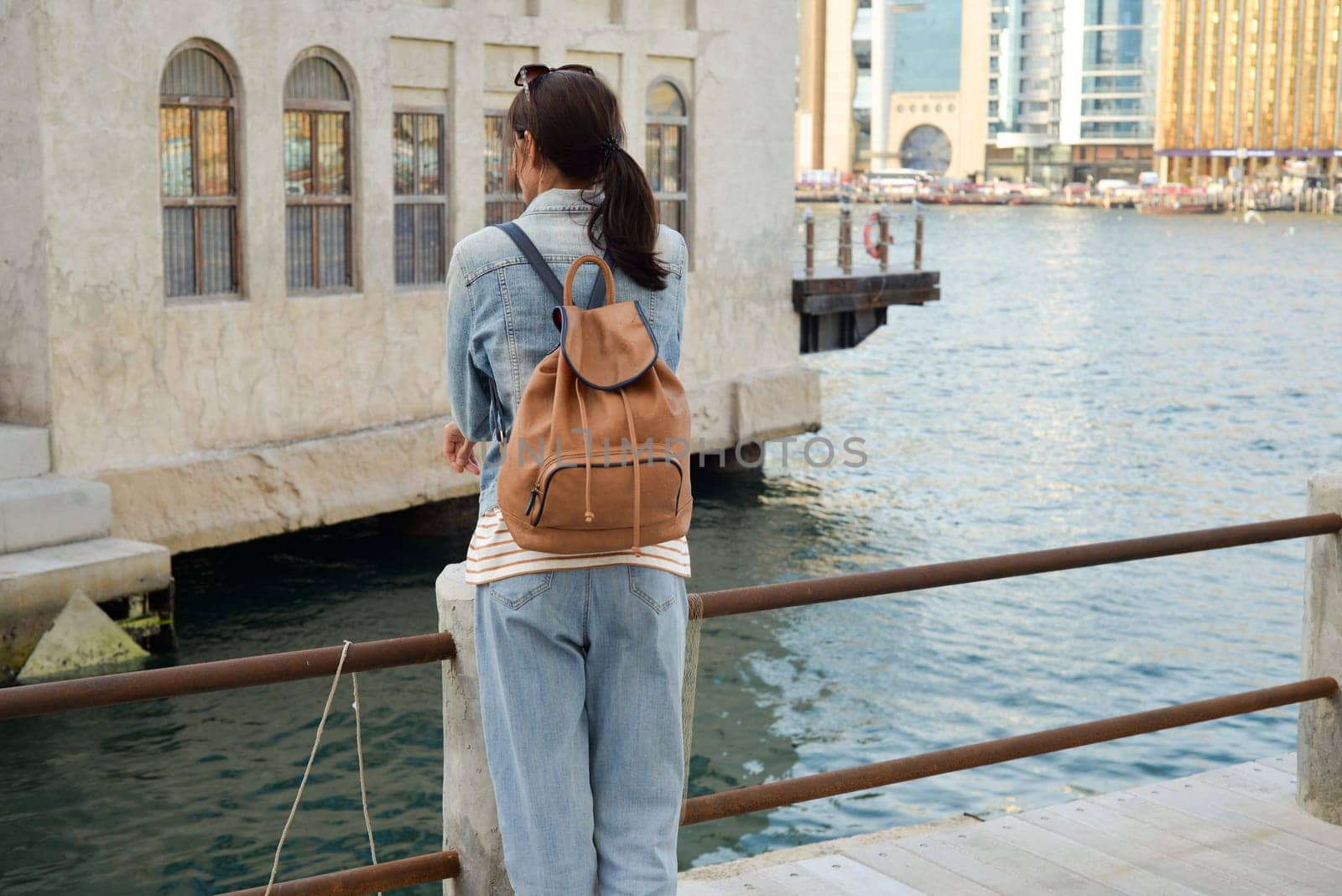A young woman with a backpack on her back admires the river in the old Dubai Creek. Back view. Journey through the Persian Gulf.