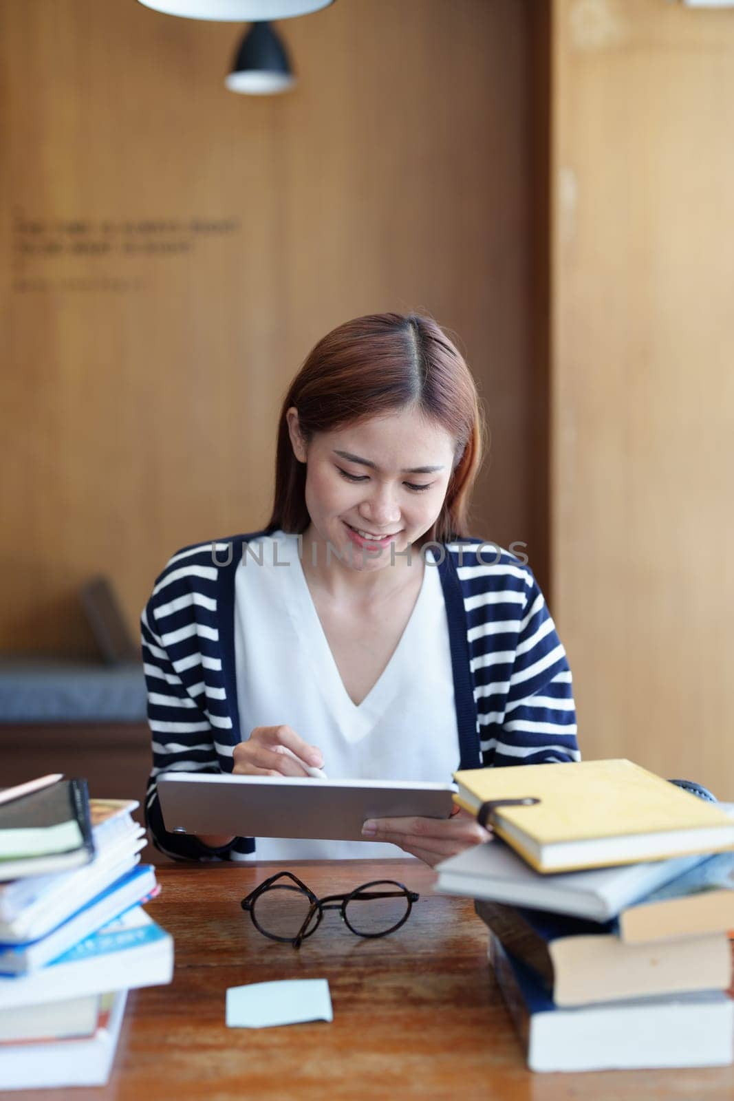 A portrait of a young Asian woman with a smiling face using a tablet computer during an online video conferencing class in a library by Manastrong