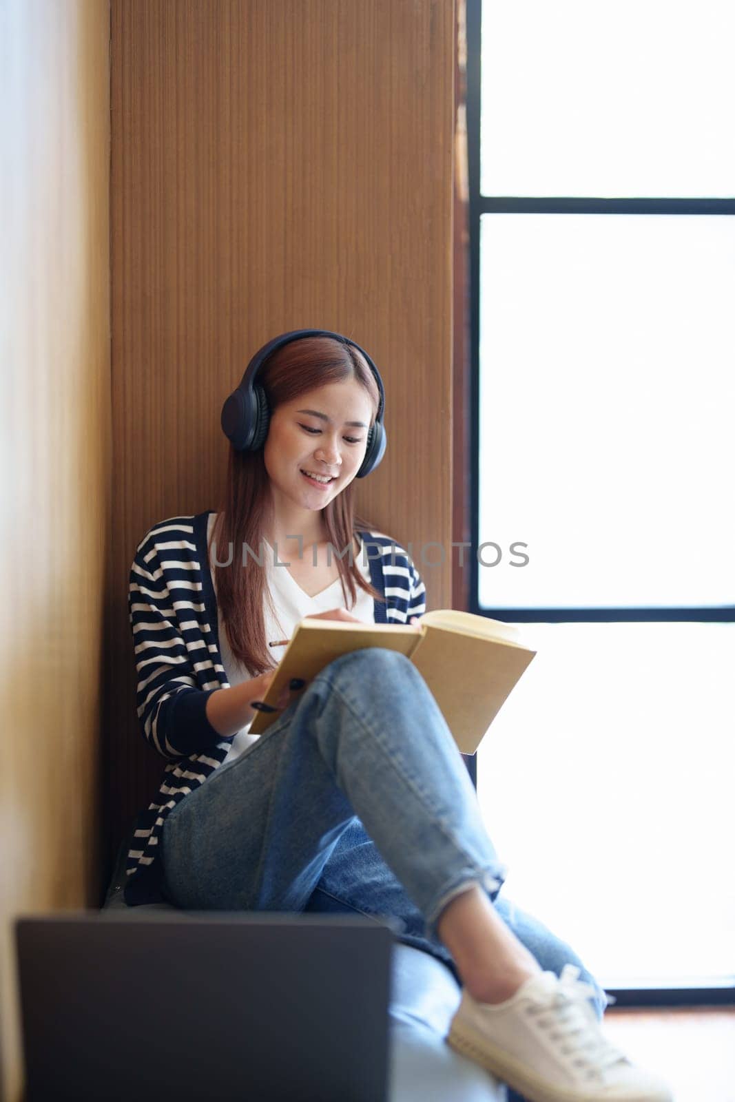 Portrait of a young Asian teenage girl in love wearing headphones, using a notebook and computer to study online via video conferencing by Manastrong