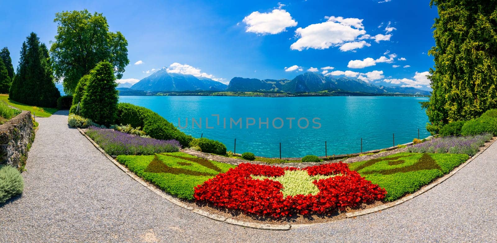 Flowerbed of the Swiss flag with boat cruise on the Thun lake and Alps mountains, Oberhofen, Switzerland. Swiss flag made of flowers and passenger cruise boat, Lake Thun, Switzerland.