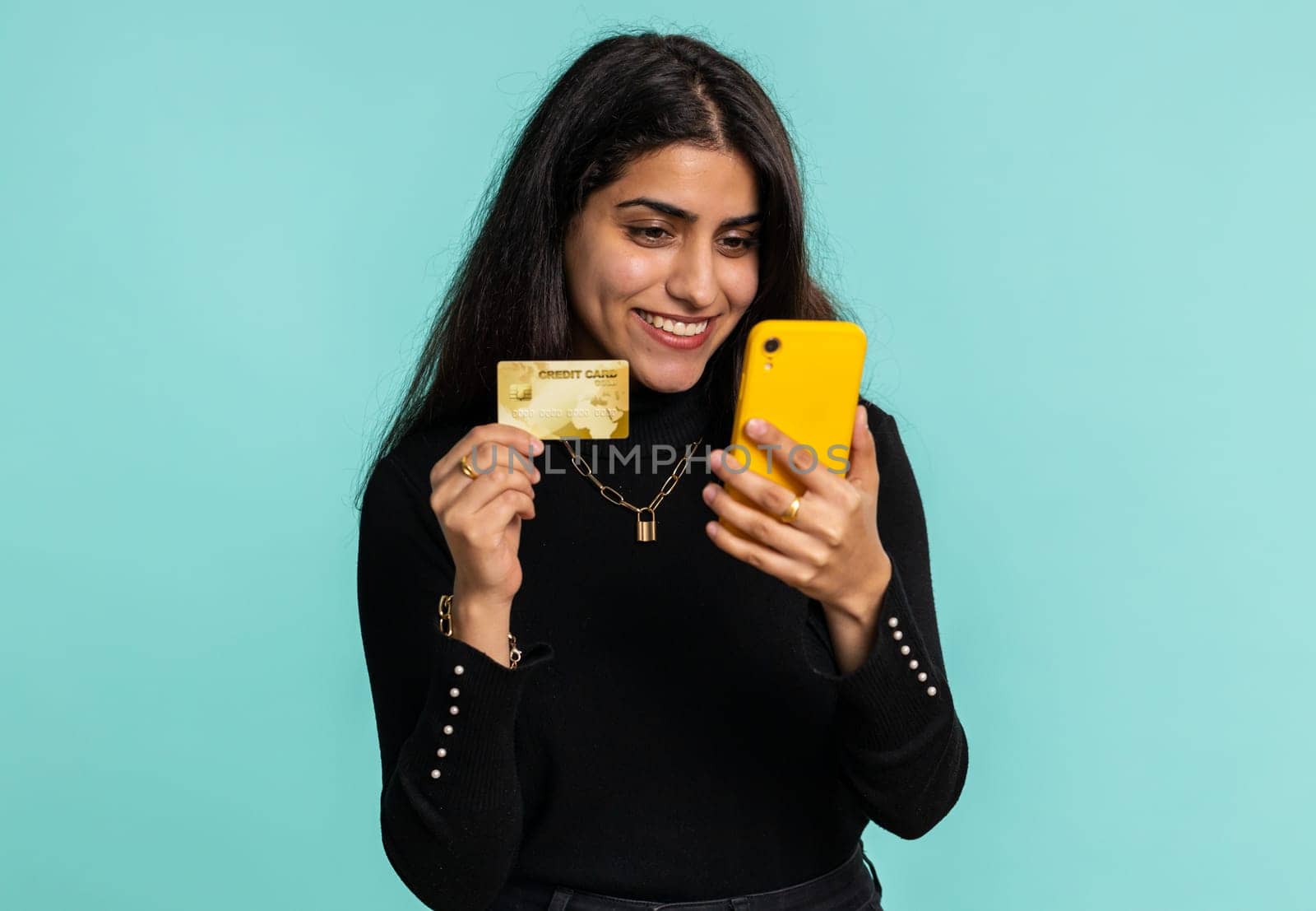 Sincere indian woman customer using credit bank card and smartphone while transferring money, purchases online shopping, payment. Finance and internet. Pretty girl isolated alone on blue background