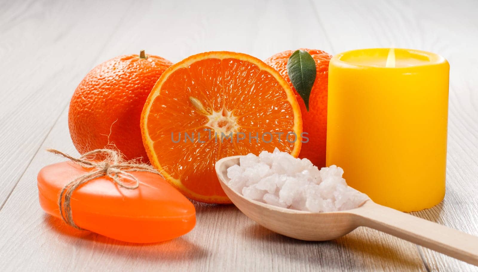 Cut orange with two whole oranges, soap, wooden spoon with white sea salt and burning yellow candle on wooden desk. Spa products and accessories.