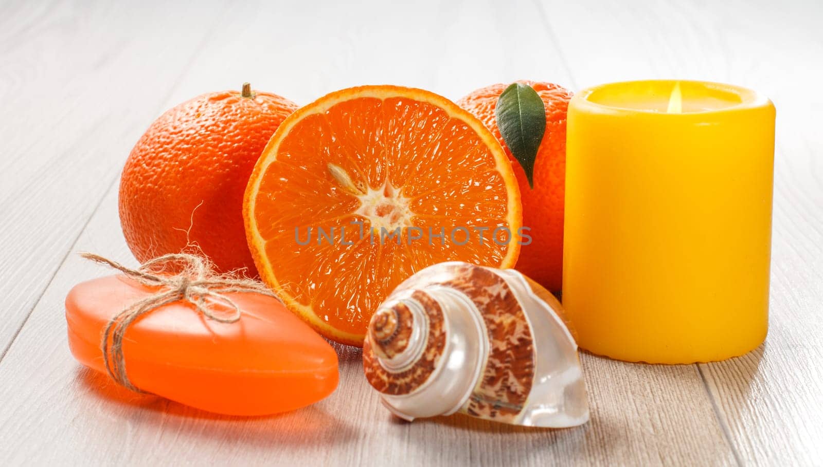 Cut orange with two whole oranges, soap, sea shell and burning candle on wooden desk. Spa products and accessories.