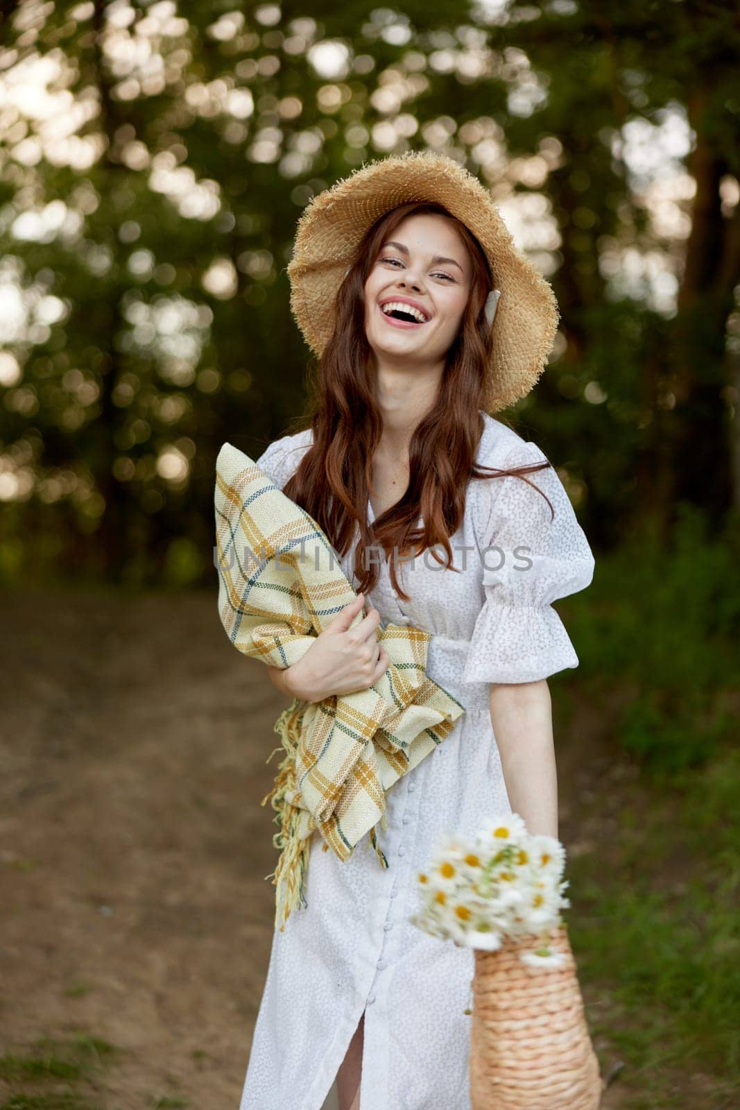 a happy woman in a light summer dress stands in nature with a wicker hat, a plaid and a basket with daisies. High quality photo