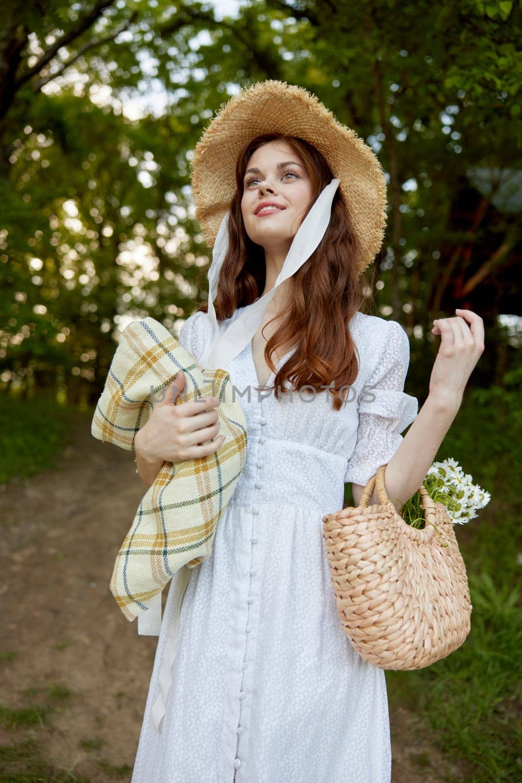 a happy woman in a light summer dress stands in nature with a wicker hat, a plaid and a basket with daisies. High quality photo