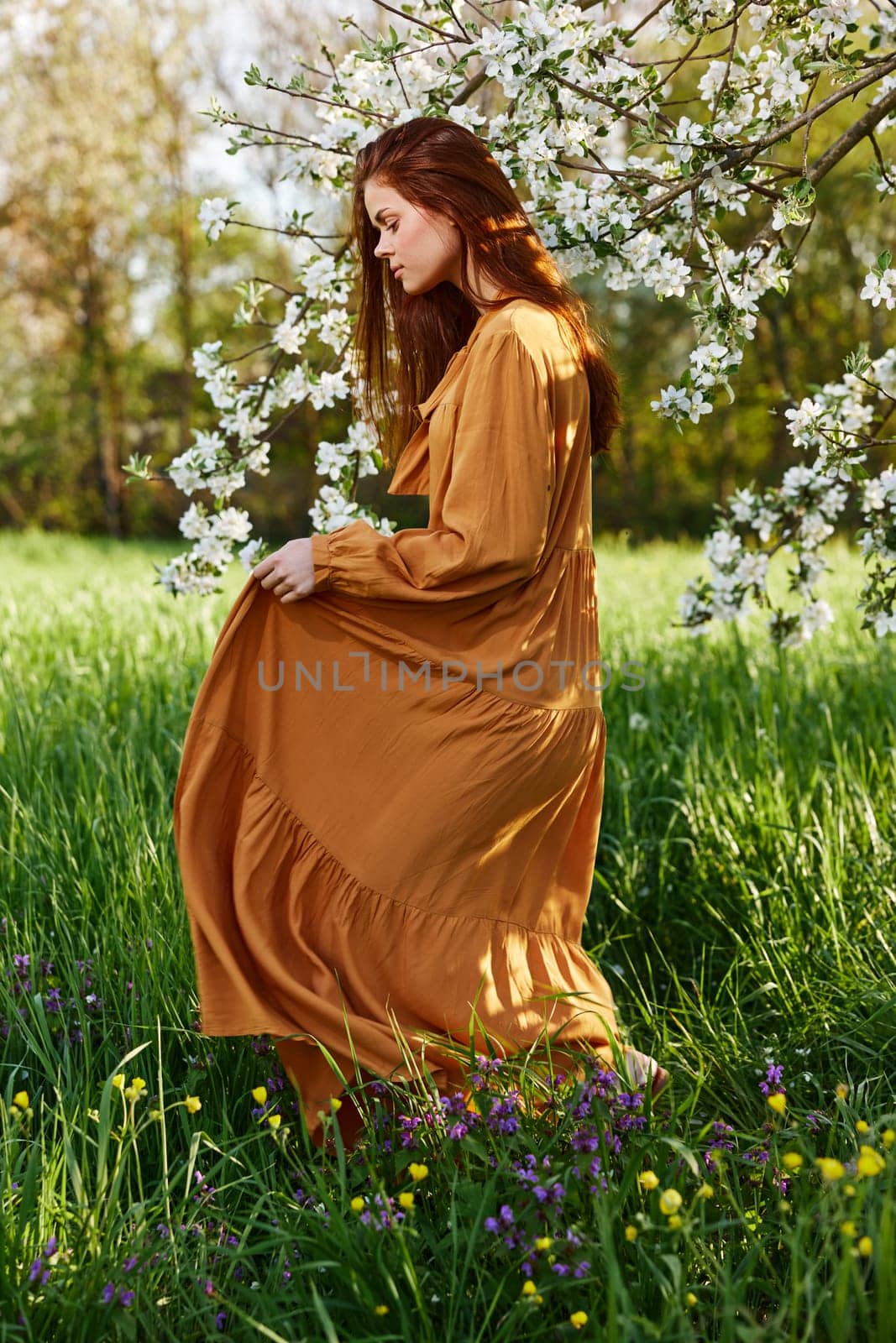 a happy, slender, sweet woman stands in a long orange dress in the tall grass near a flowering tree and happily smiling throws up the hem of her dress, looking away. High quality photo